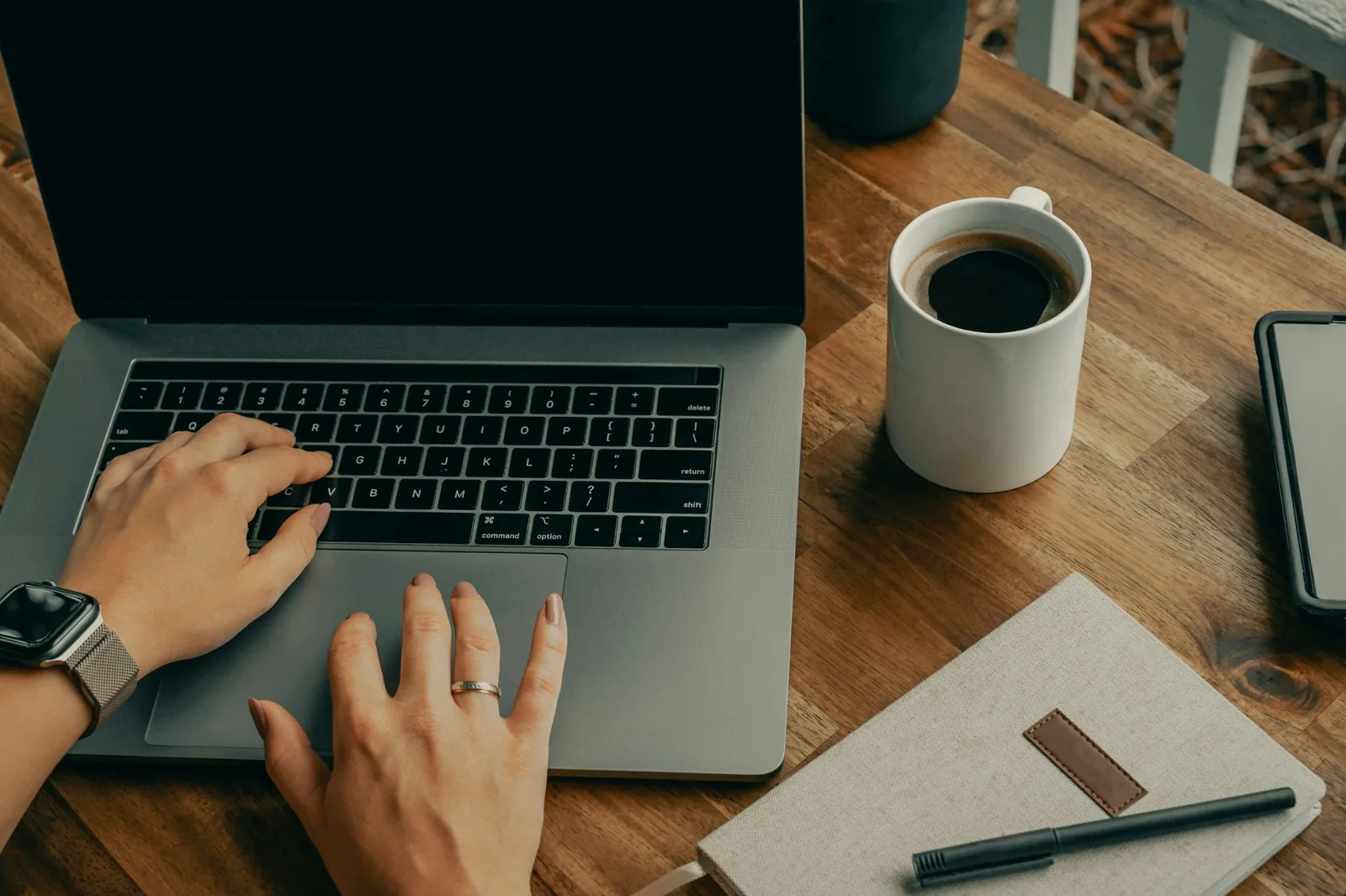 Ahşap masada laptop, kahve kupası, defter ve kalemle çalışan eller