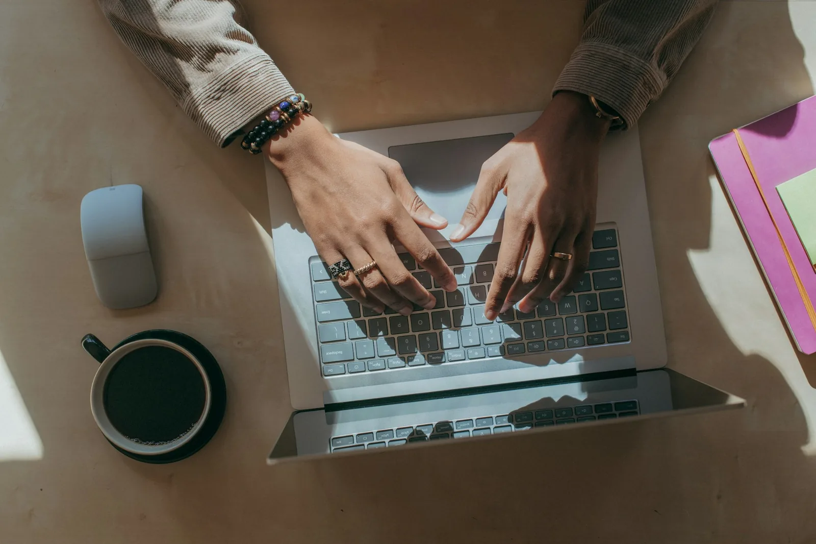 Güneş ışığı alan bir masada laptopta yazan eller, yanında kahve ve defter