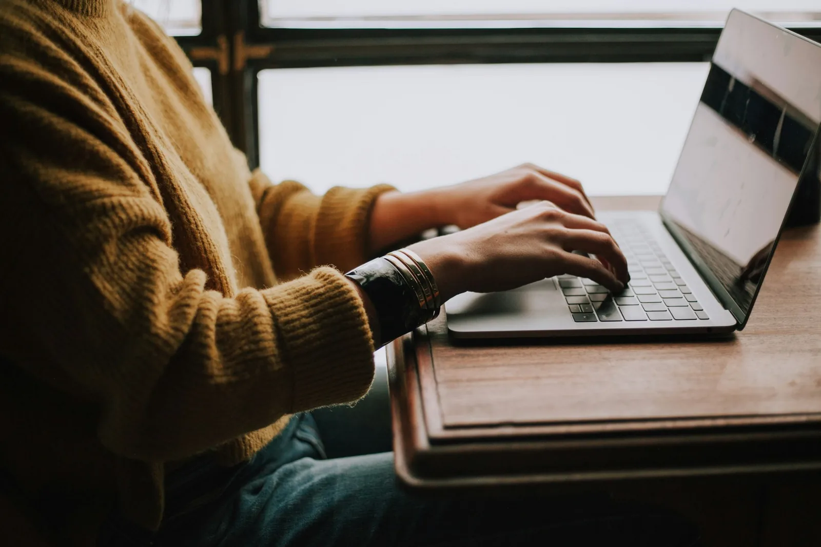 Pencere önünde hardal renkli kazağıyla laptopta yazan bir öğrenci